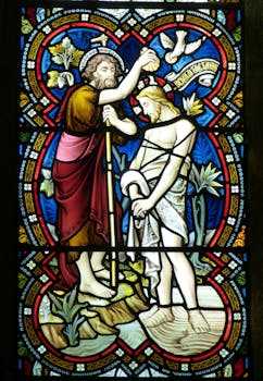 Detailed stained glass window depicting a religious baptism scene with vibrant colors.